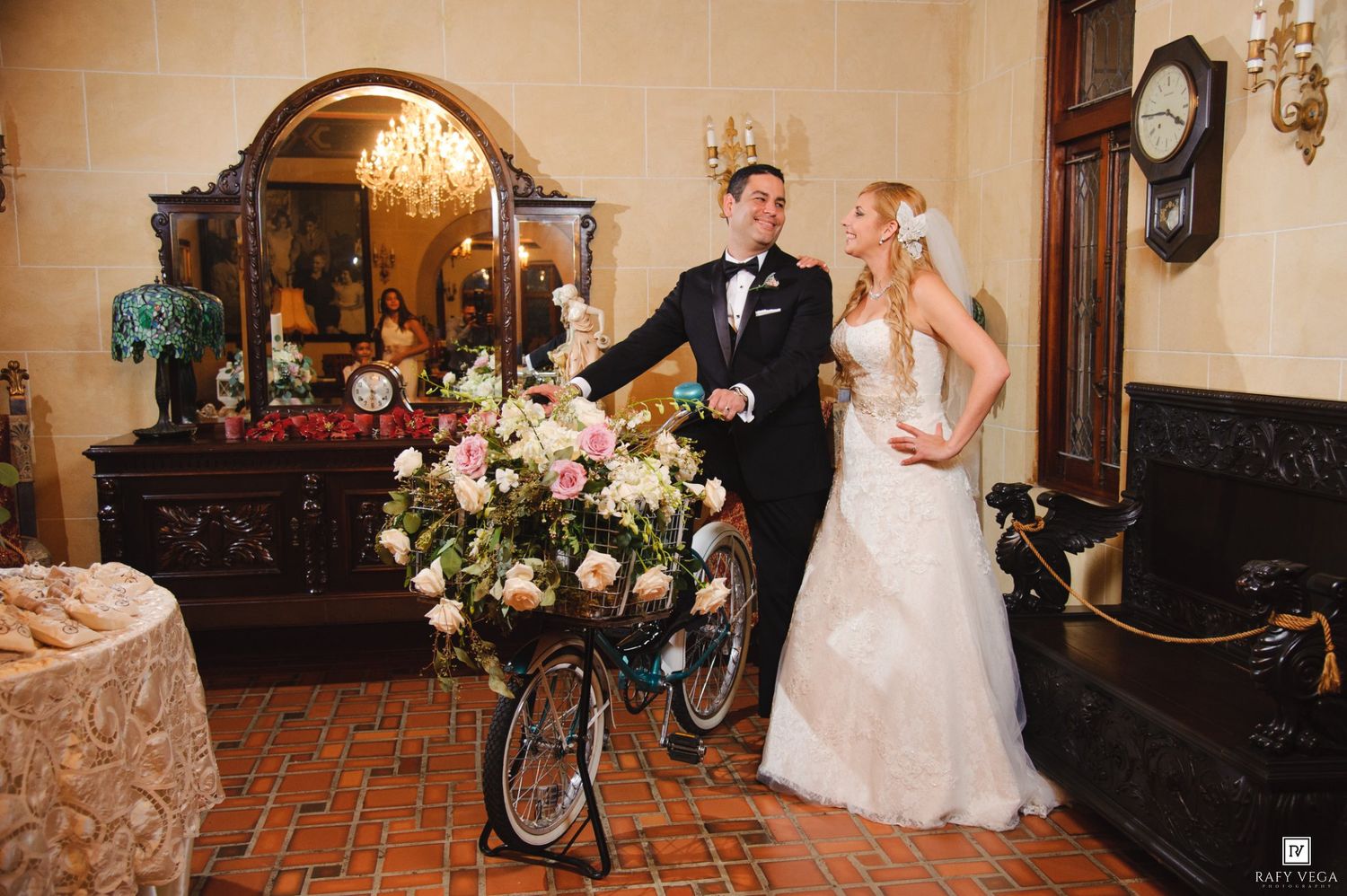 Bride and groom pose with a floral-decorated bicycle inside an elegant venue with antique furniture and warm lighting.