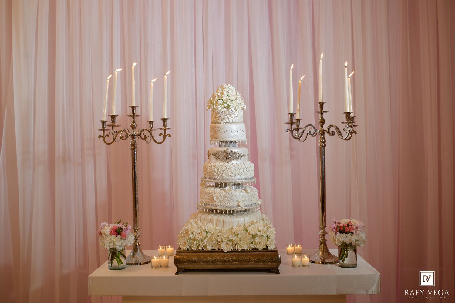 Elegant multi-tiered white wedding cake flanked by tall candelabras with lit candles on a decorated table with pink backdrop.