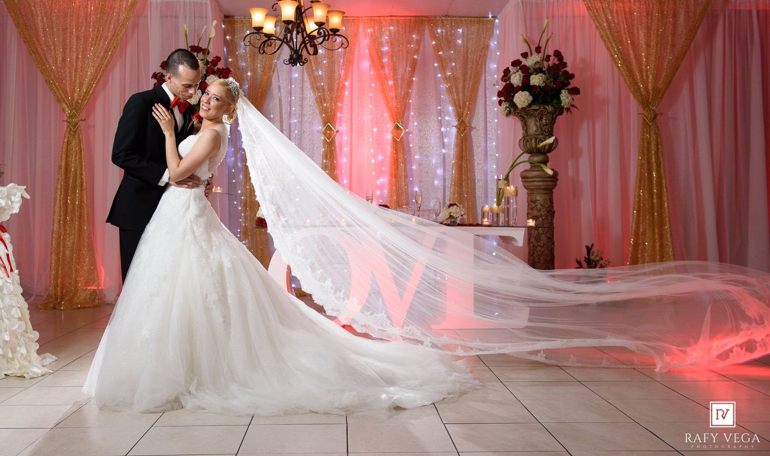Bride and groom share romantic kiss at elegant wedding venue with pink draping, chandelier, and flowing cathedral veil.
