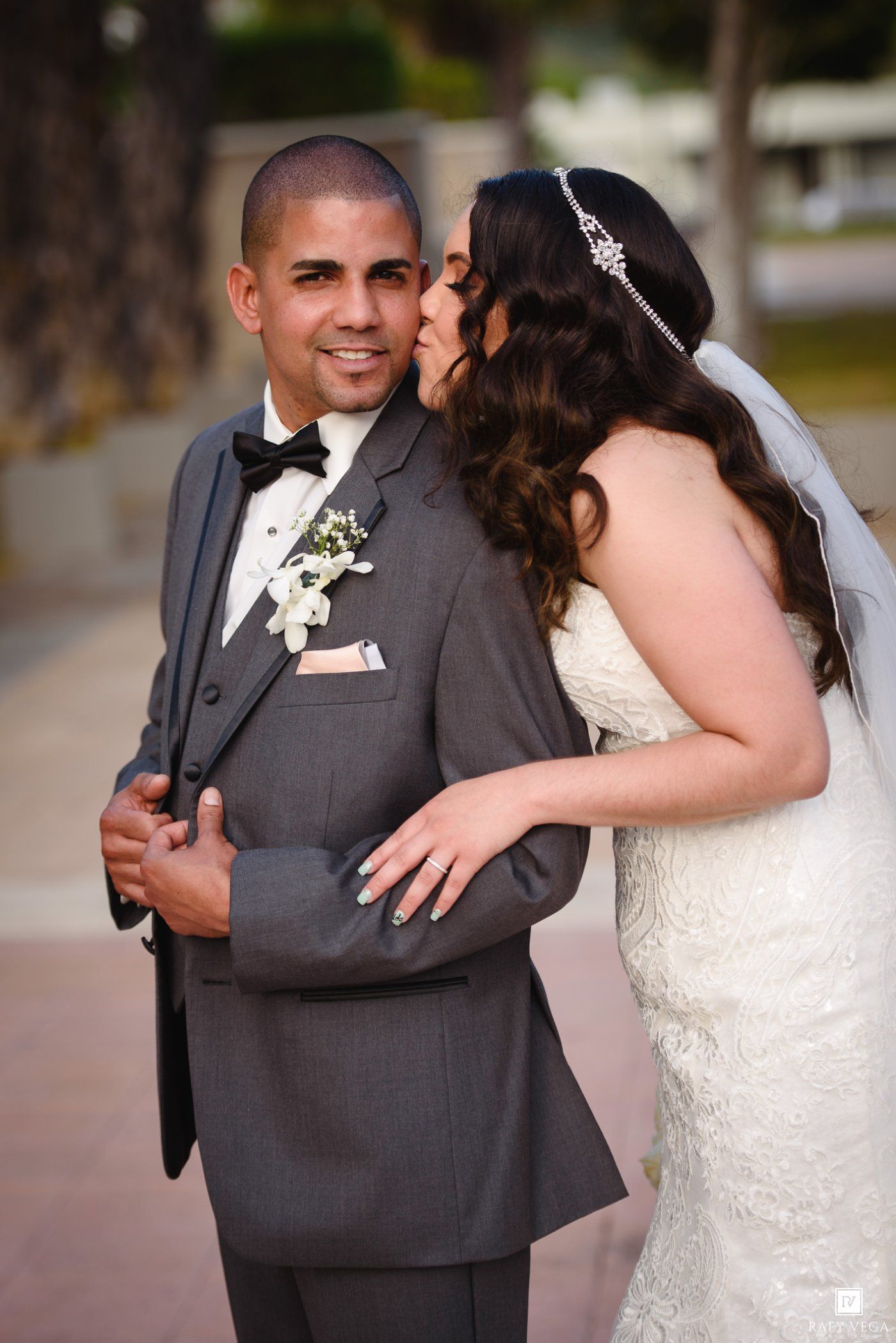 Bride in white lace gown kisses groom in gray tuxedo on cheek outdoors at wedding celebration.