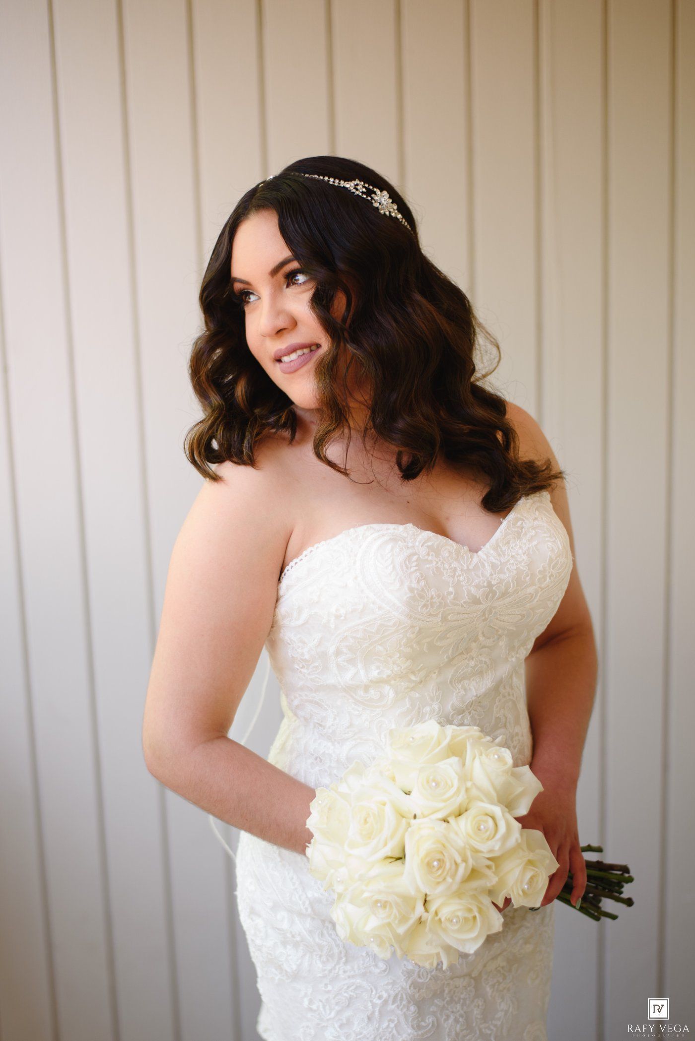 Bride in strapless lace wedding gown holding white rose bouquet, wearing crystal tiara, posing elegantly indoors.
