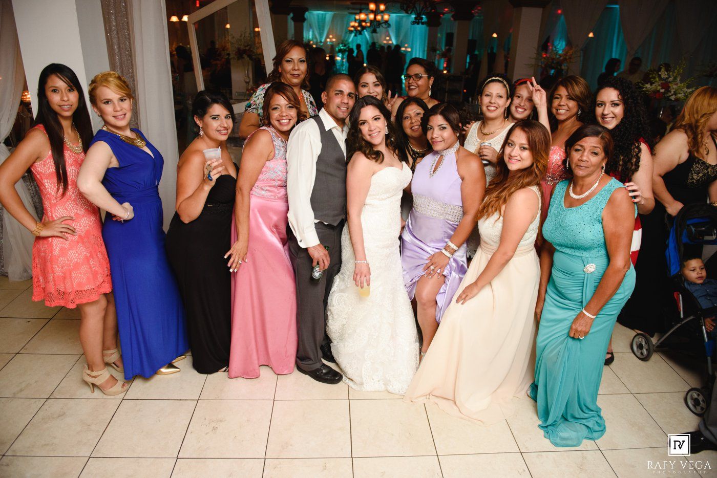 Bride and groom posing with large group of wedding guests in colorful formal attire on dance floor.