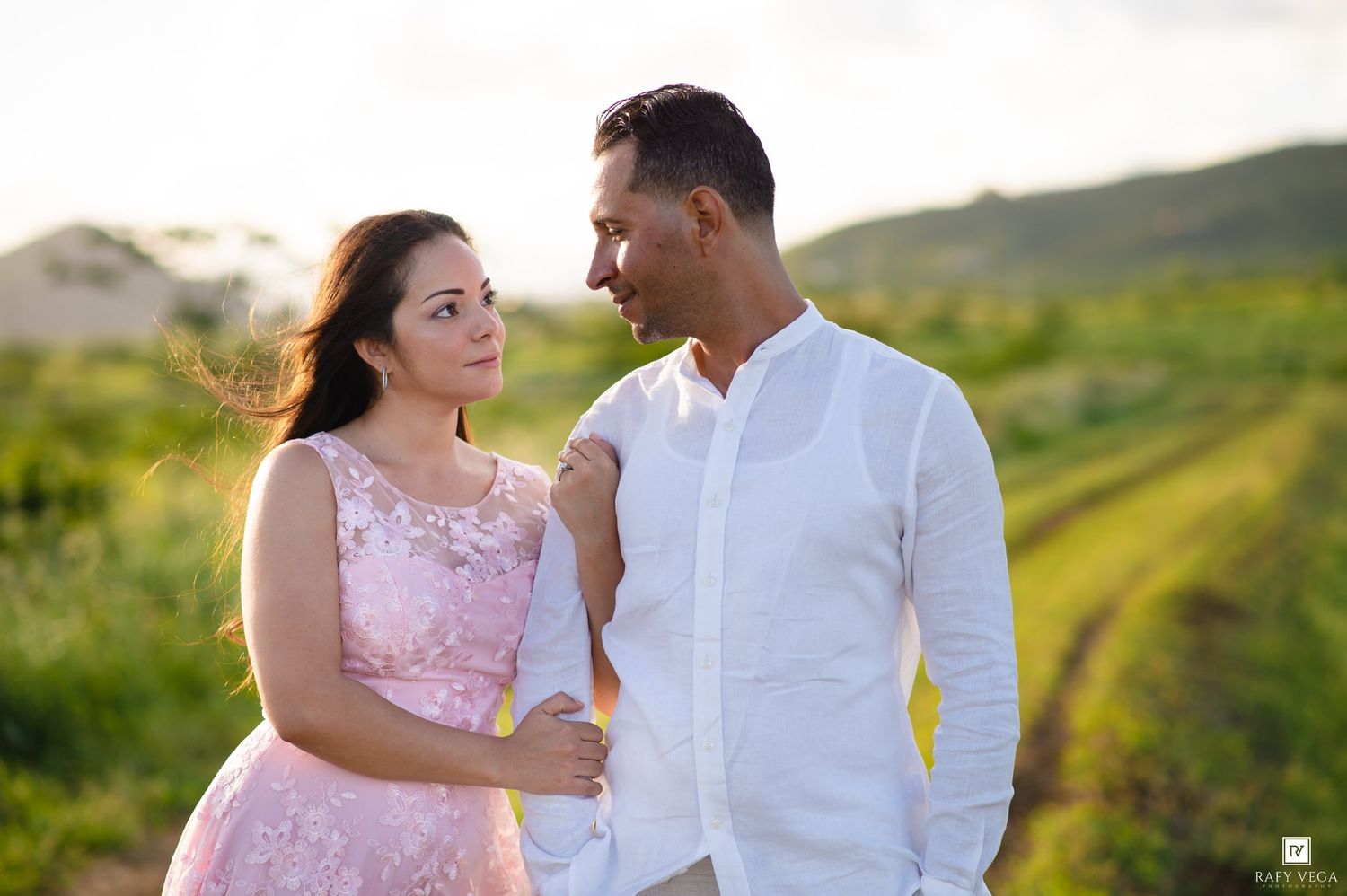 Couple in romantic outdoor photoshoot, woman in pink lace dress and man in white shirt, green hills backdrop.