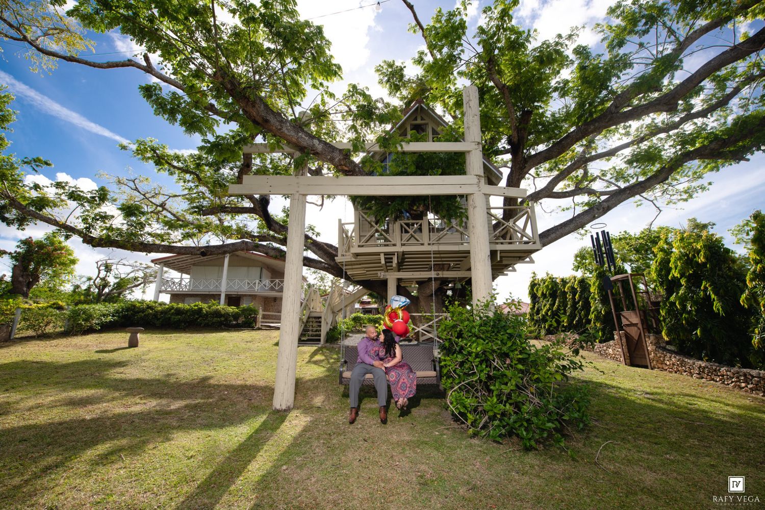 Unique treehouse structure built around a large tree in a lush garden, with visitors relaxing on benches below.