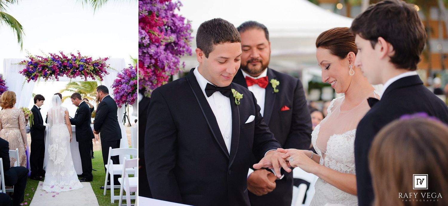 Bride and groom exchange rings during outdoor wedding ceremony with purple floral arch and guests in background.