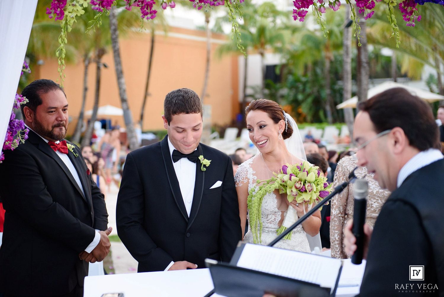 Bride and groom exchange vows at outdoor wedding ceremony under floral arch with officiant and witness present.