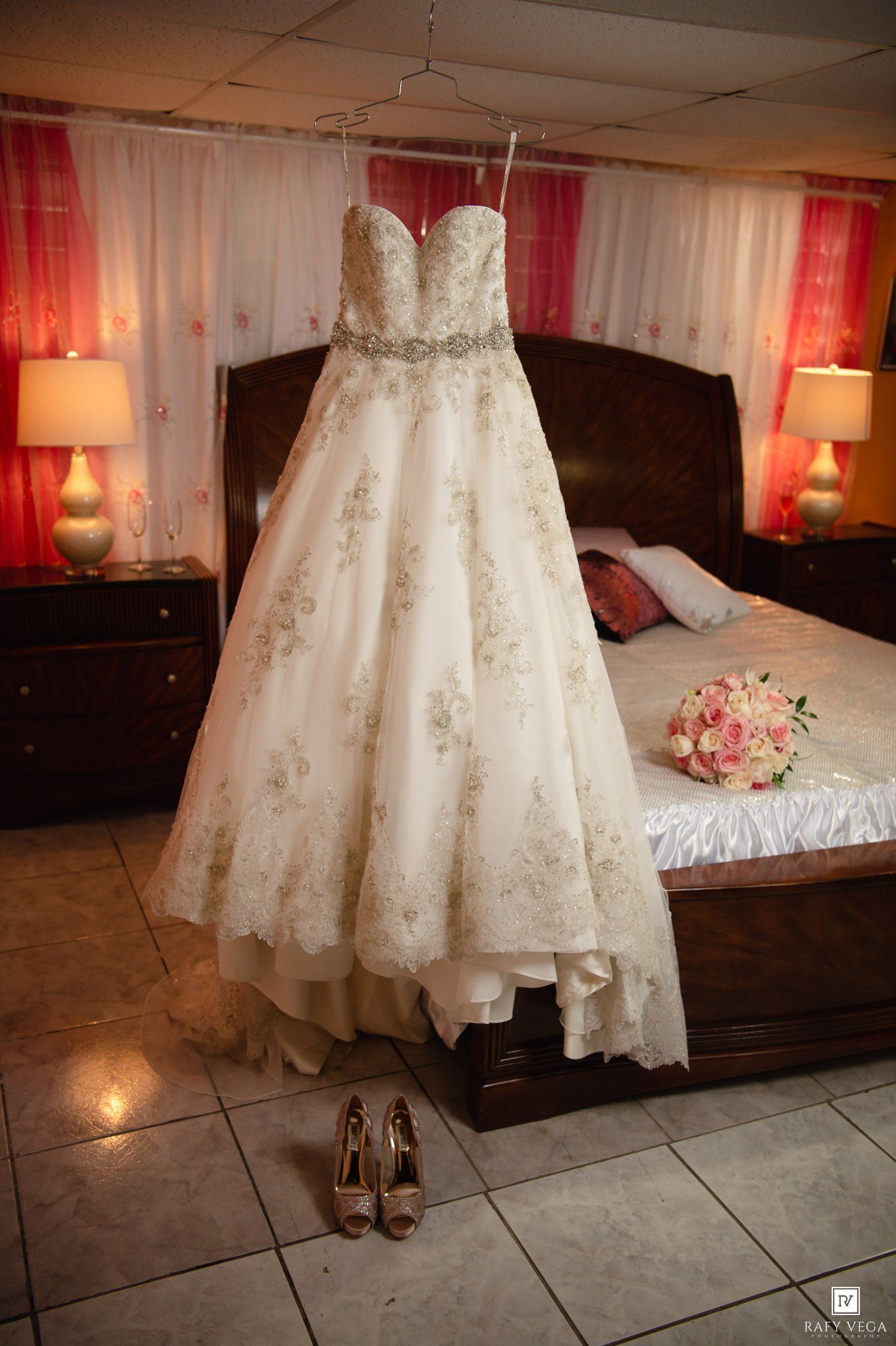 Elegant strapless white wedding gown with floral embroidery hanging in a bedroom with heels and bouquet.