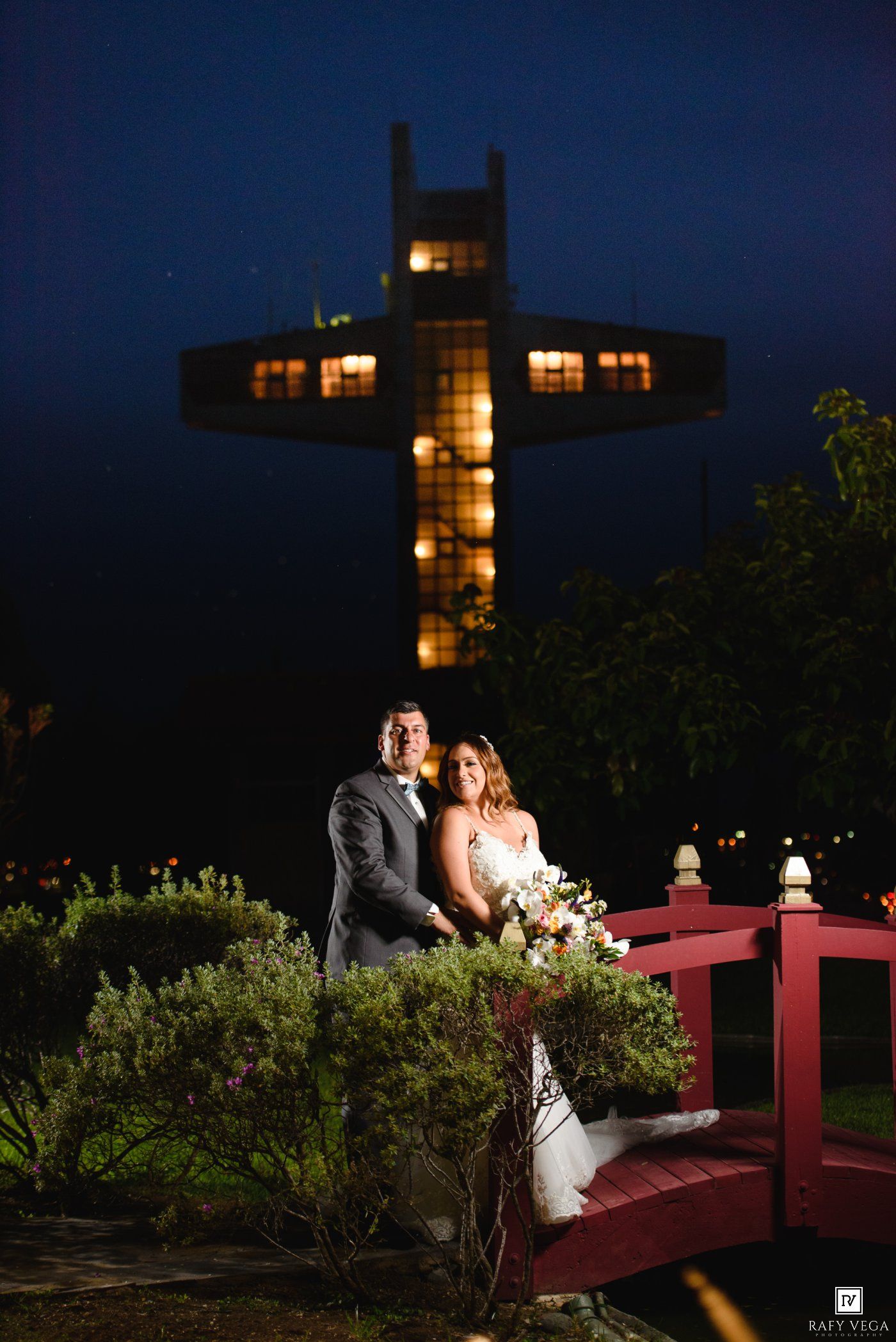 Bride and groom pose on a red bridge at night with an illuminated cross landmark glowing behind them.