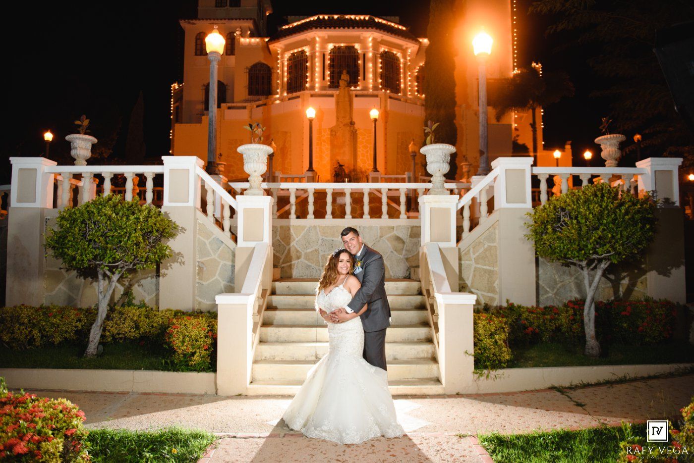 Bride and groom embrace on elegant white staircase outside illuminated wedding venue at night.
