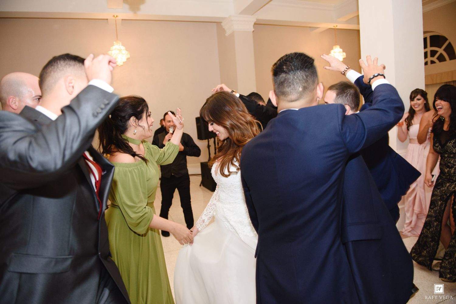 Bride and groom dancing with guests in a circle at a wedding reception in an elegant white venue.