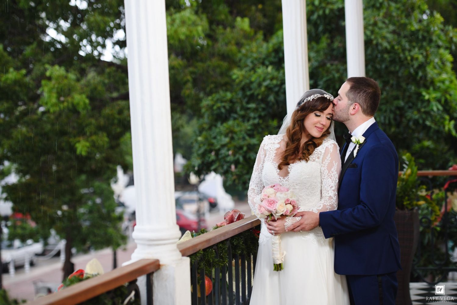 Bride and groom share a romantic moment on a white columned porch, bride holding a pink bouquet in a lace wedding dress.