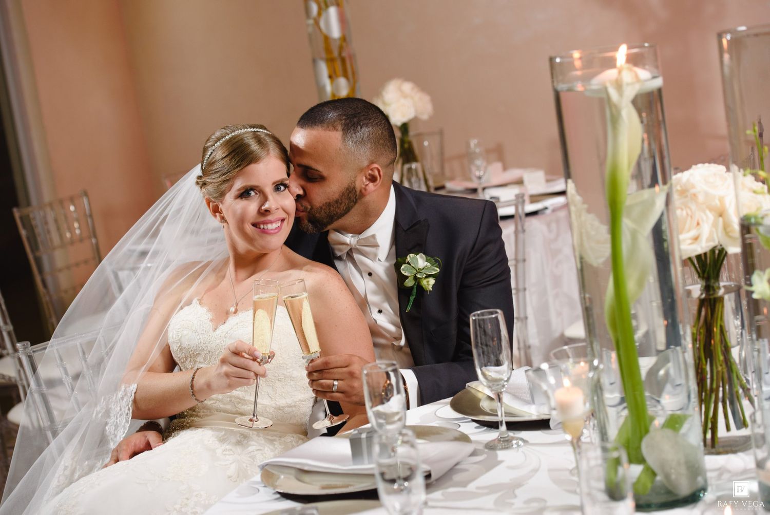 Bride and groom share a romantic moment at wedding reception table, holding champagne flutes with floral centerpieces nearby.