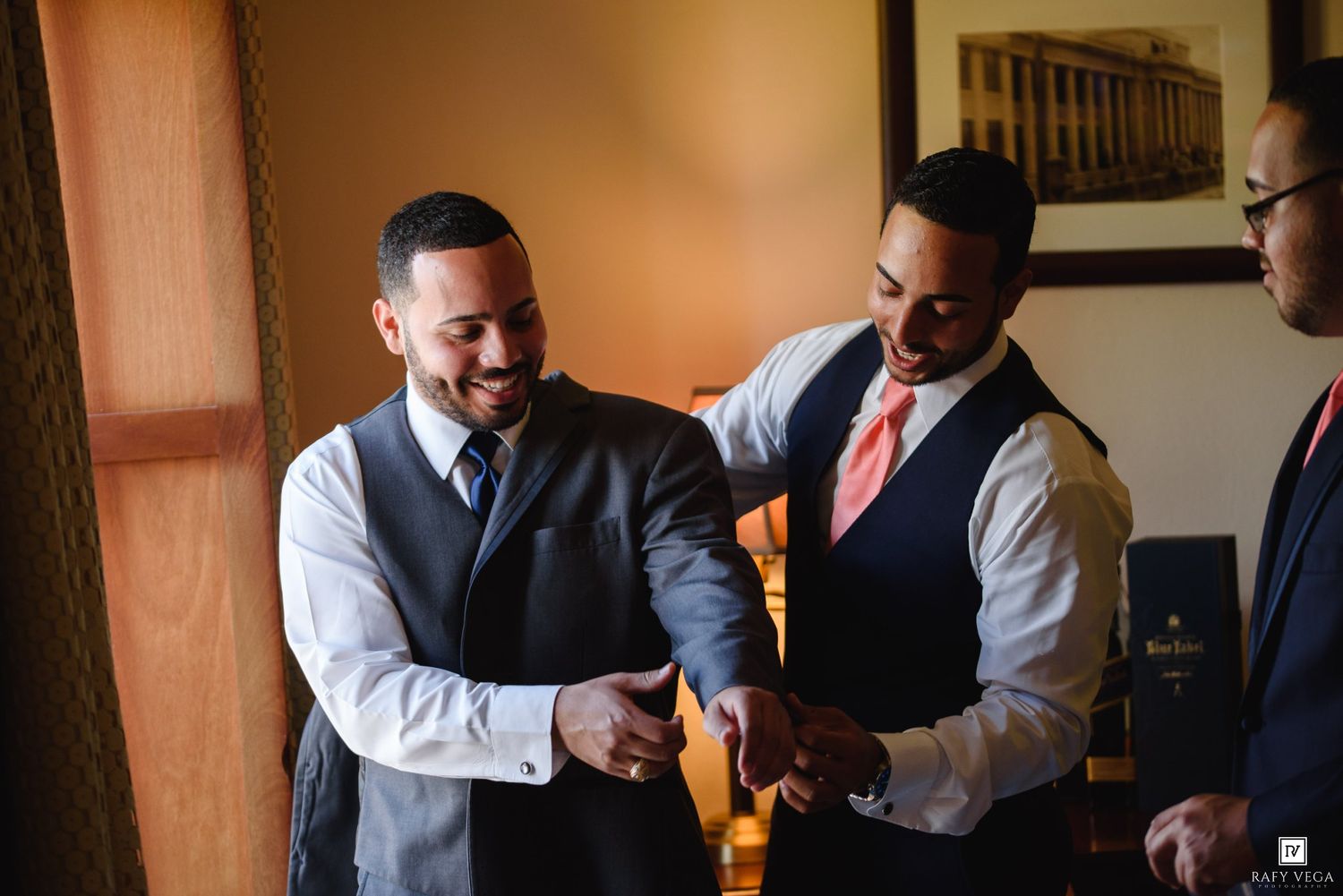 Two groomsmen in vests help a groom with his suit jacket before a wedding ceremony.