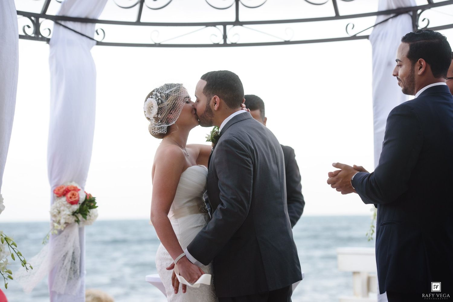 Bride and groom share first kiss at outdoor beach wedding ceremony under white draped arch with ocean backdrop.