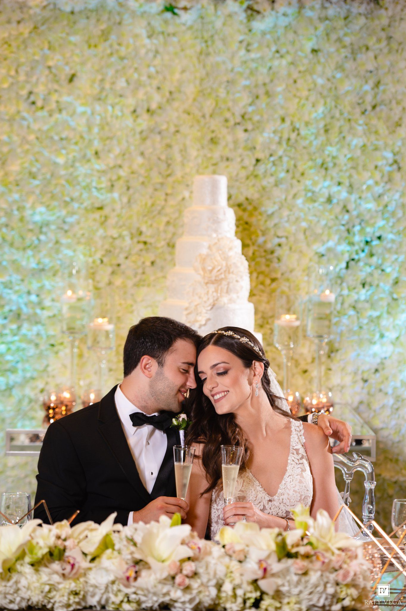 Bride and groom share an intimate moment at sweetheart table with white floral wall and tiered wedding cake backdrop.