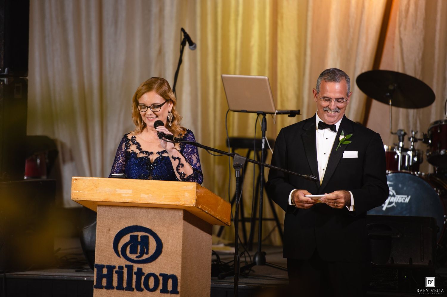 Woman speaking at Hilton podium beside man in tuxedo at formal gala event.