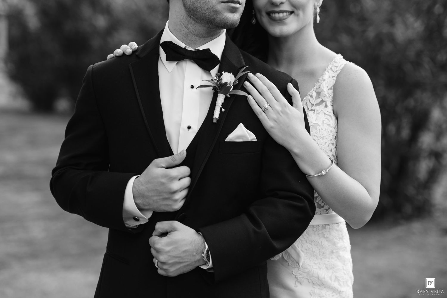 Black and white close-up of bride embracing groom in tuxedo, showcasing wedding rings and boutonniere.