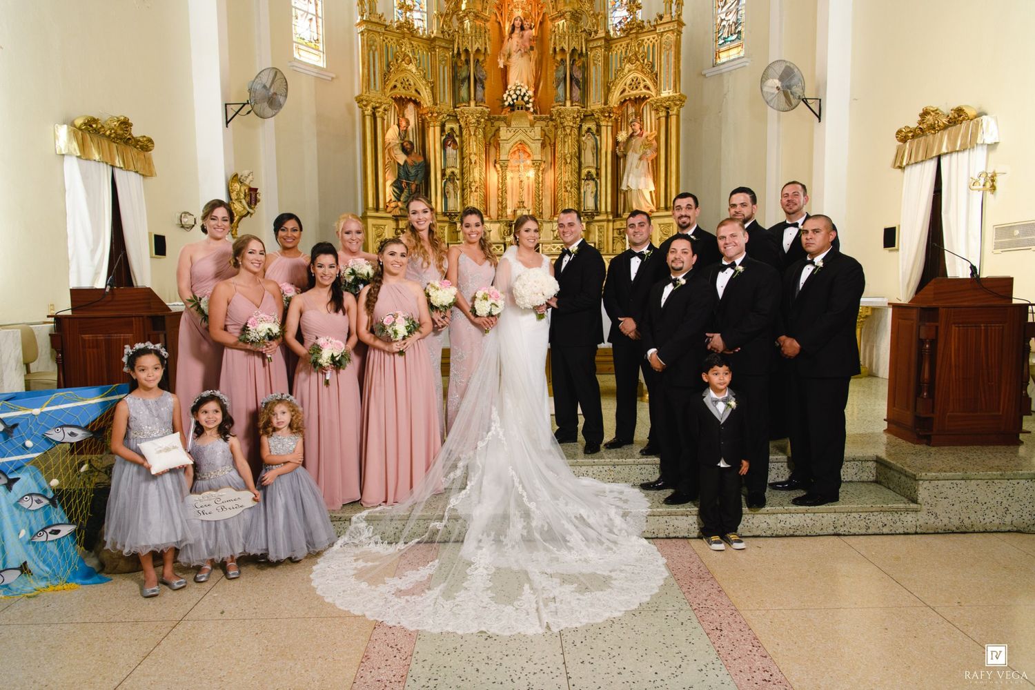 Wedding party poses in a church with bridesmaids in pink dresses, groomsmen in tuxedos, and bride in a lace gown.
