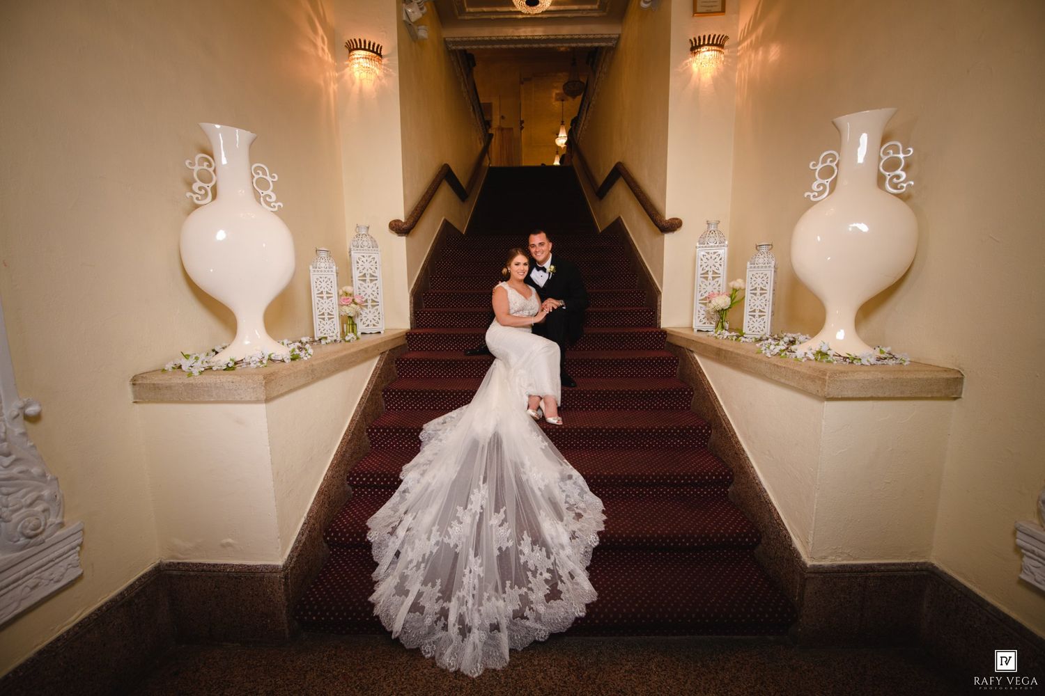 Bride and groom embrace on elegant staircase with white vases and floral decor, bride wearing lace mermaid gown.