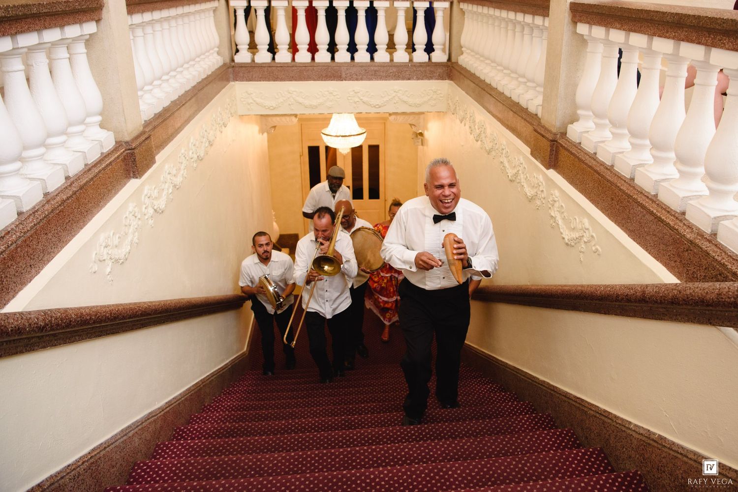 Jazz band musicians descending a grand staircase with red carpet, playing brass instruments in formal attire.