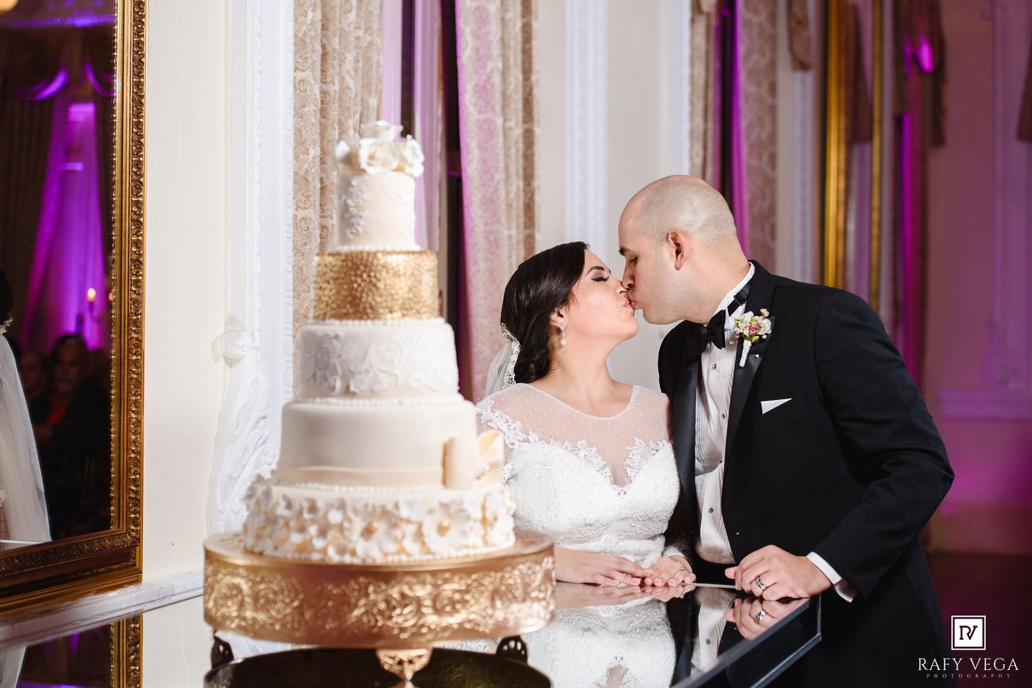 Bride and groom share a kiss beside a tall white and gold tiered wedding cake at an elegant reception venue.