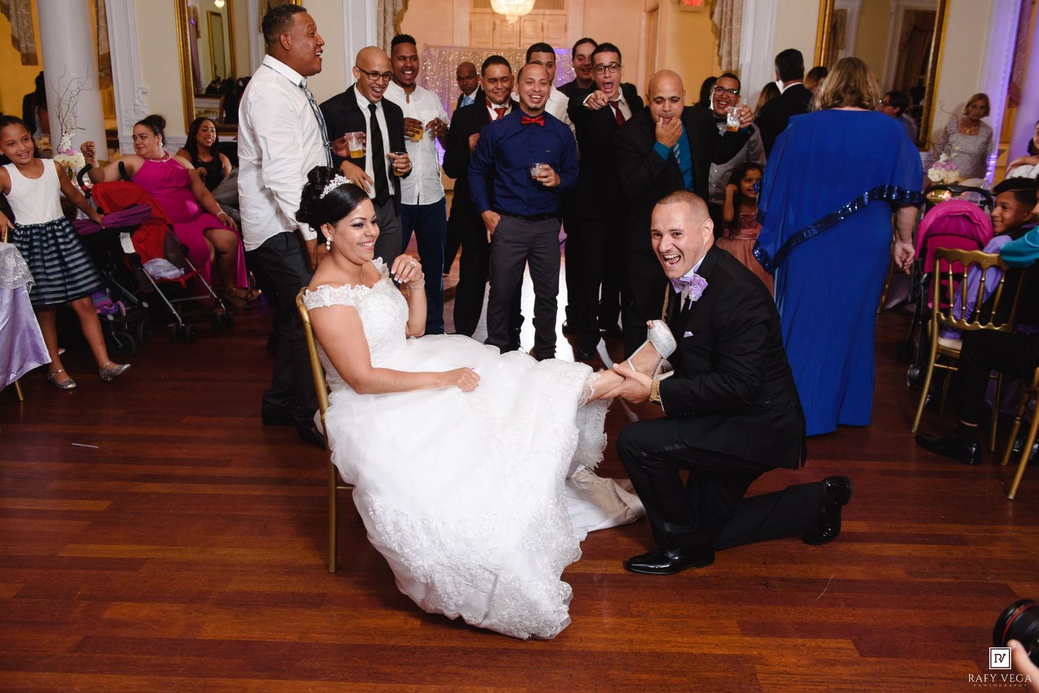 Groom kneeling before bride during garter removal at elegant wedding reception with guests watching and applauding.