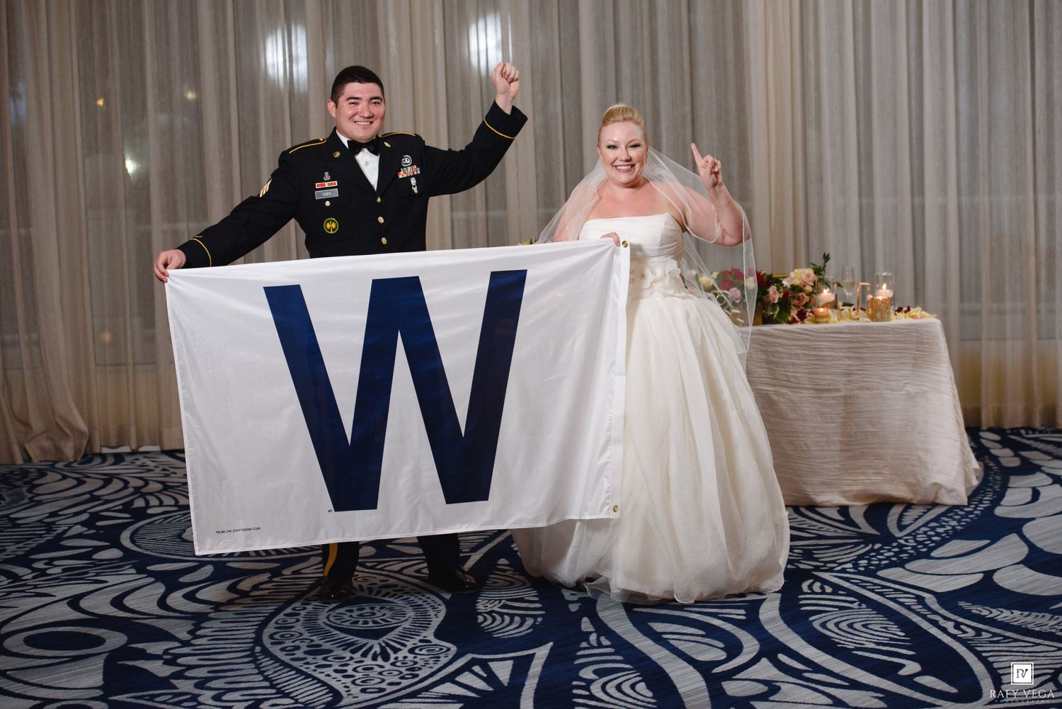 Newlywed couple in wedding attire celebrating while holding a Chicago Cubs W flag at their wedding reception.