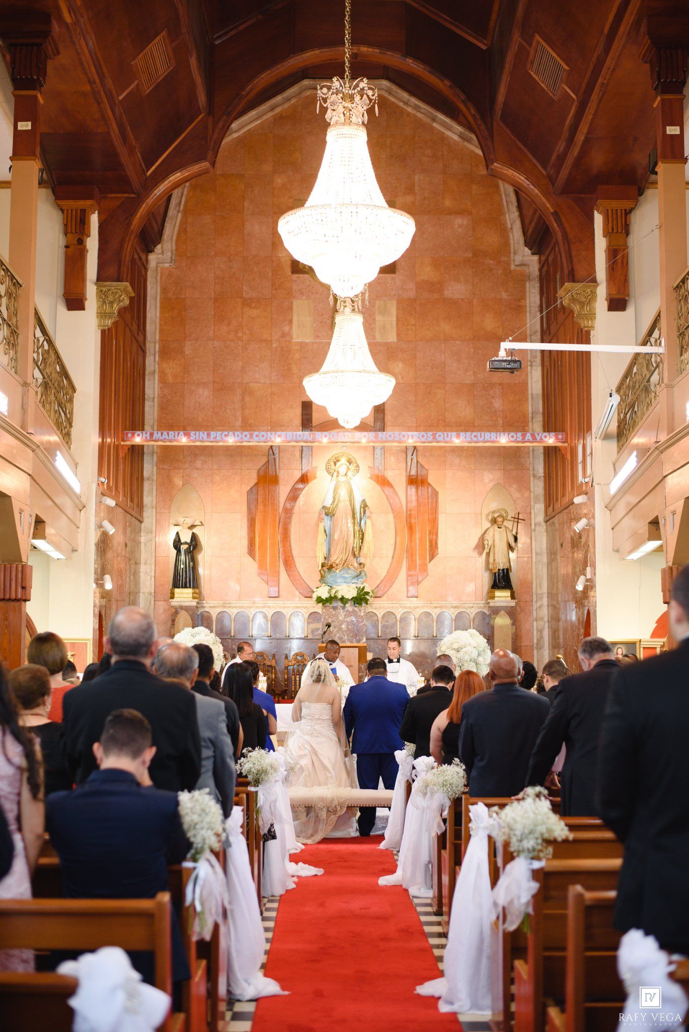 Elegant church wedding ceremony with bride and groom at altar, red carpet aisle, white floral pew decorations, and crystal chandelier.