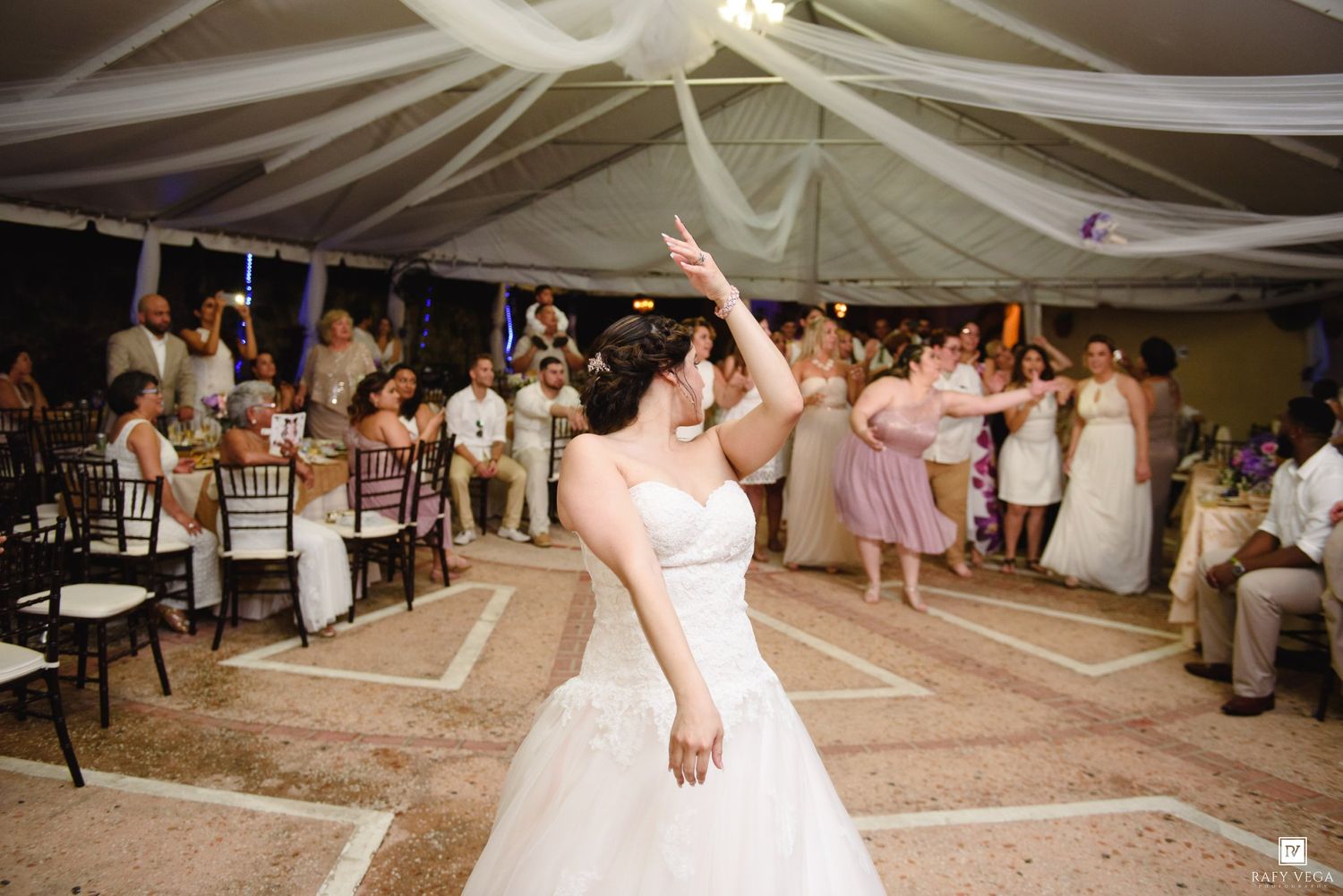 Bride tossing bouquet to excited guests at an outdoor wedding reception under a white tent.