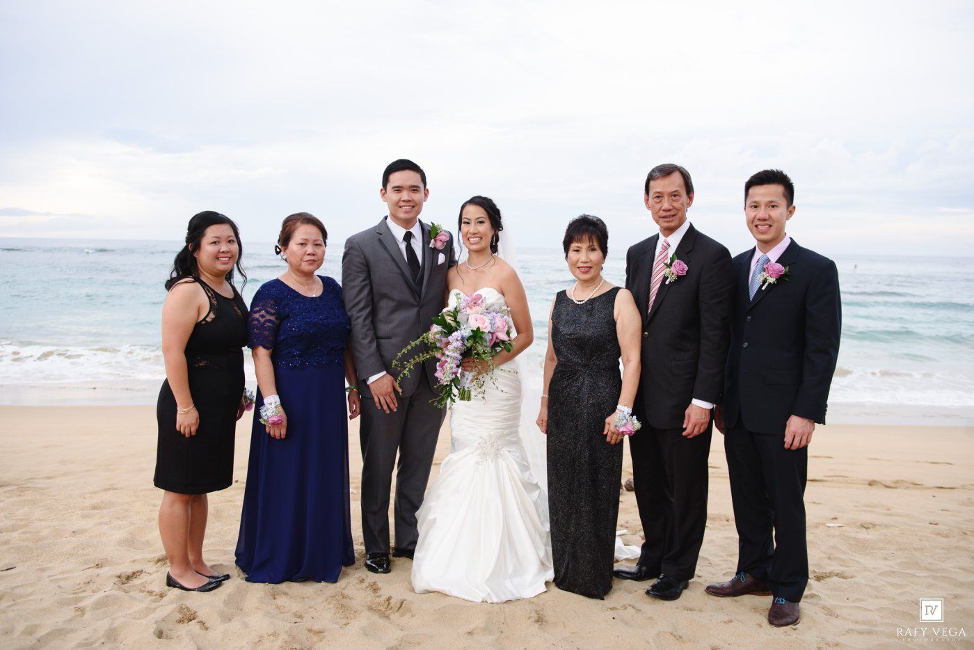 Wedding party posing on a beach, bride in white gown with groom and family members dressed in formal attire by the ocean.