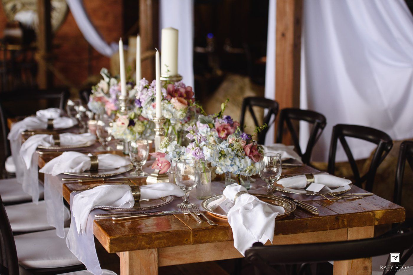 Elegant rustic wedding reception table with floral centerpieces, white linens, gold candlesticks, and place settings in a barn venue.
