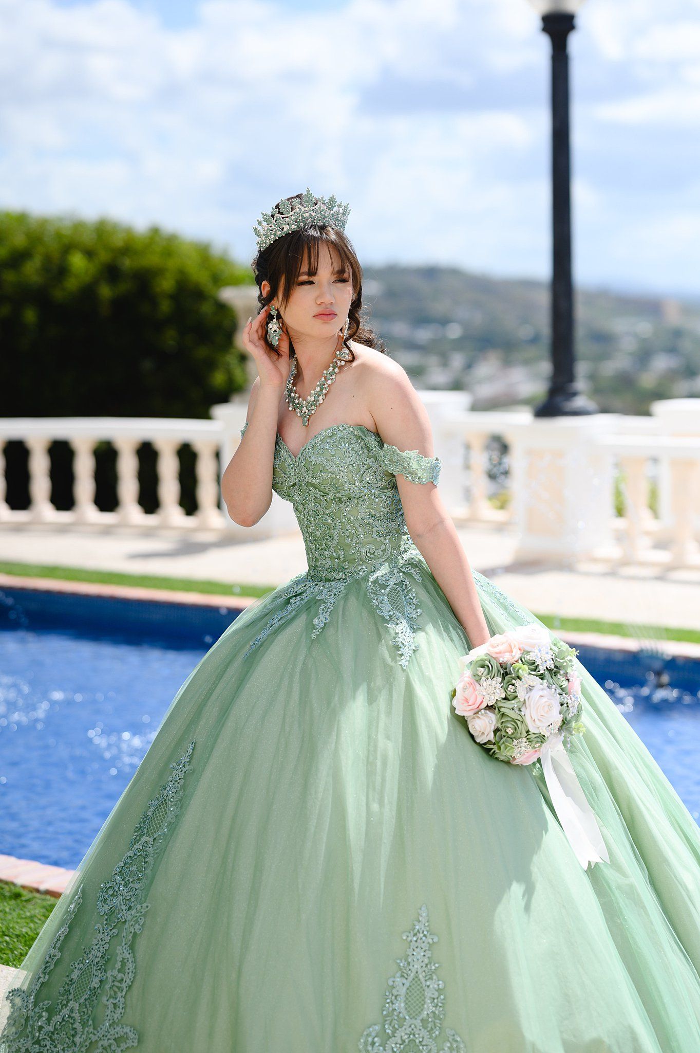 Young woman in sage green quinceañera ballgown with crown and bouquet poses by pool.