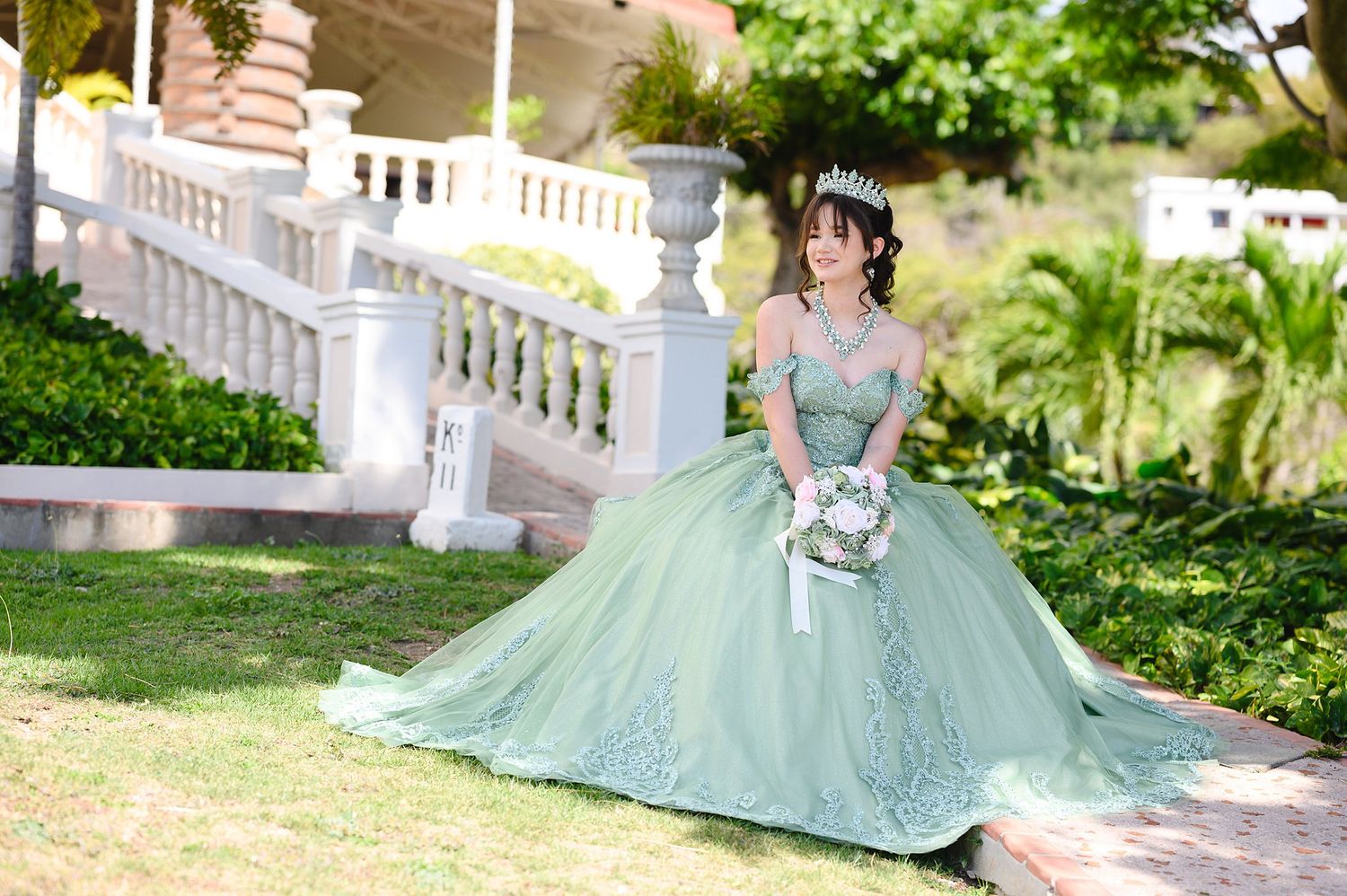 Young woman in elegant sage green quinceañera ball gown with tiara and bouquet posing outdoors.
