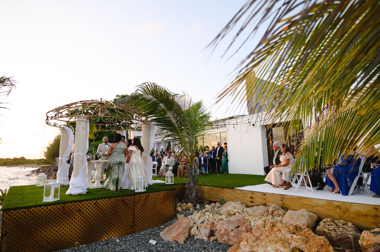 Outdoor wedding ceremony near waterfront with palm trees, white pergola, and guests gathered on green lawn at sunset at Ocean View Ponce.
