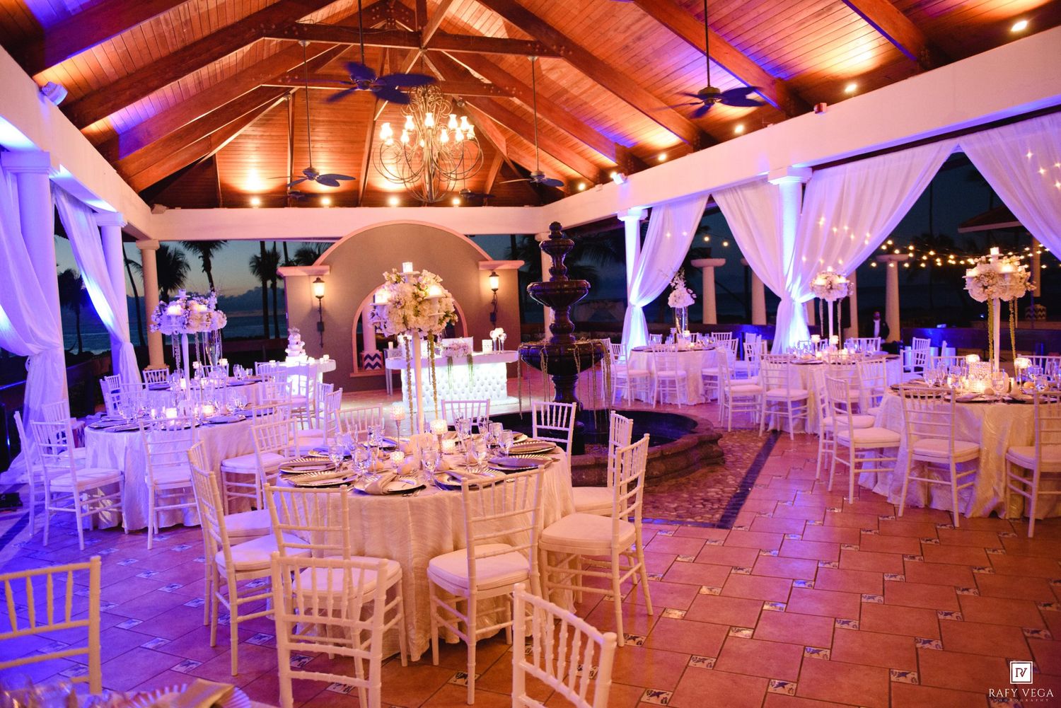 Stunning indoor wedding reception venue with white chiavari chairs, round tables, and warm amber uplighting.