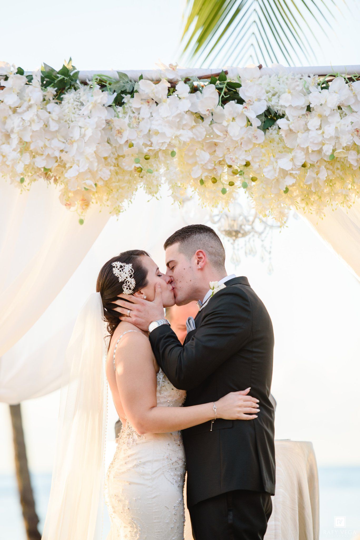 Bride and groom share first kiss under floral arch with white roses at outdoor beach wedding ceremony.
