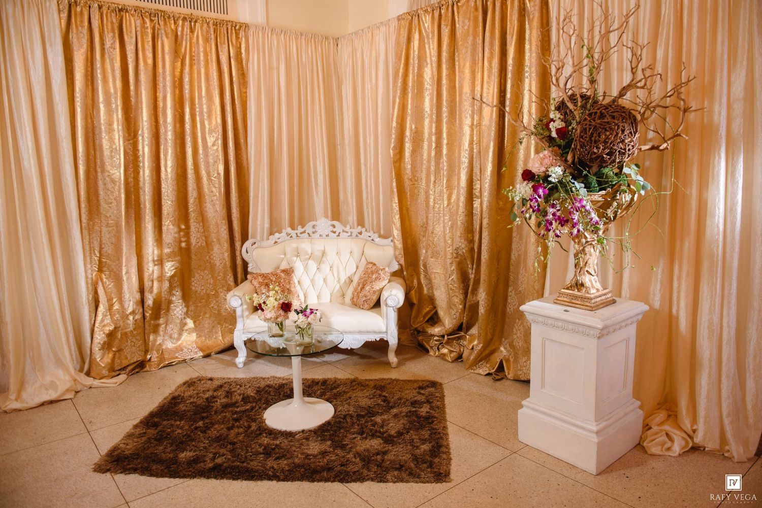 Elegant gold draped backdrop with white tufted loveseat, floral arrangement on pedestal, and brown fur rug.