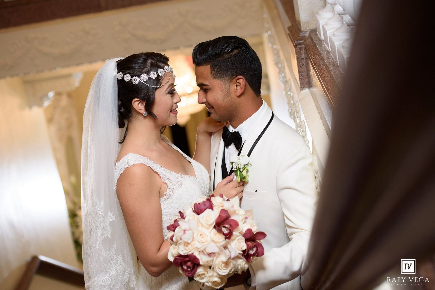 Bride and groom share a romantic moment, she holds red and white bouquet, wearing white gown and floral headpiece.