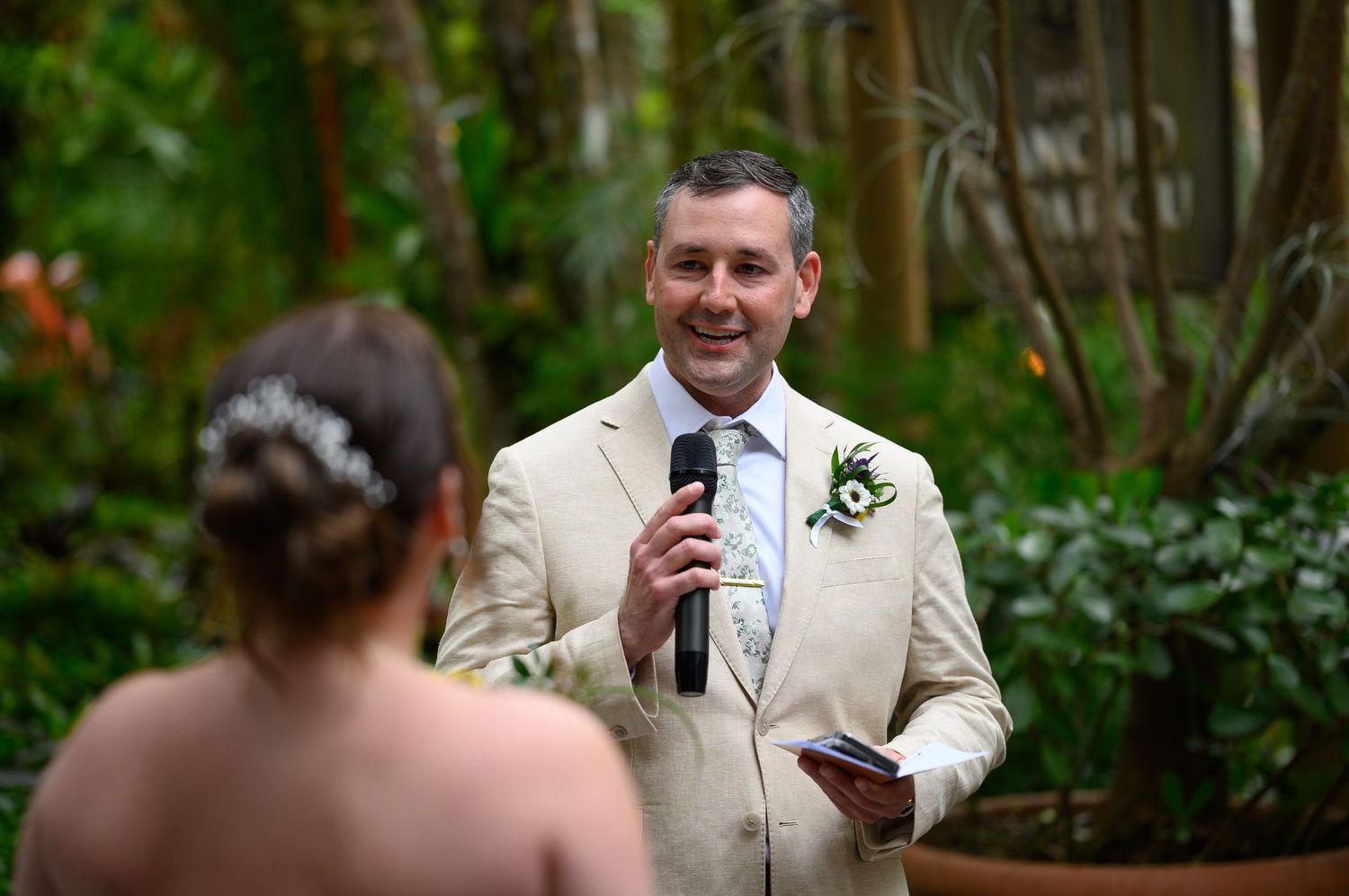 Groom in beige suit speaking vows at outdoor garden wedding ceremony, holding microphone and small book.