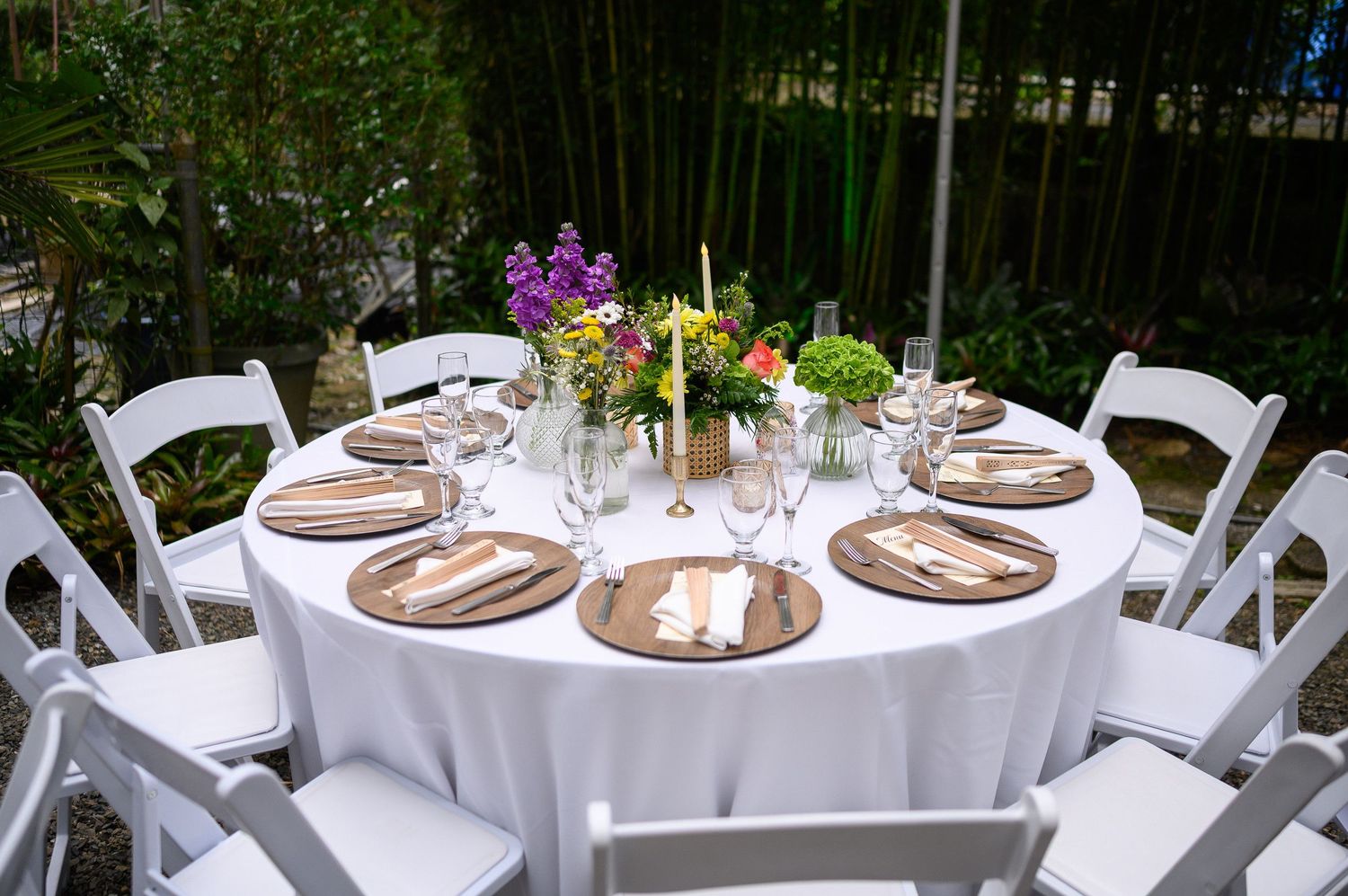 Elegant outdoor round table with white tablecloth, gold chargers, white folding chairs, and colorful floral centerpiece.