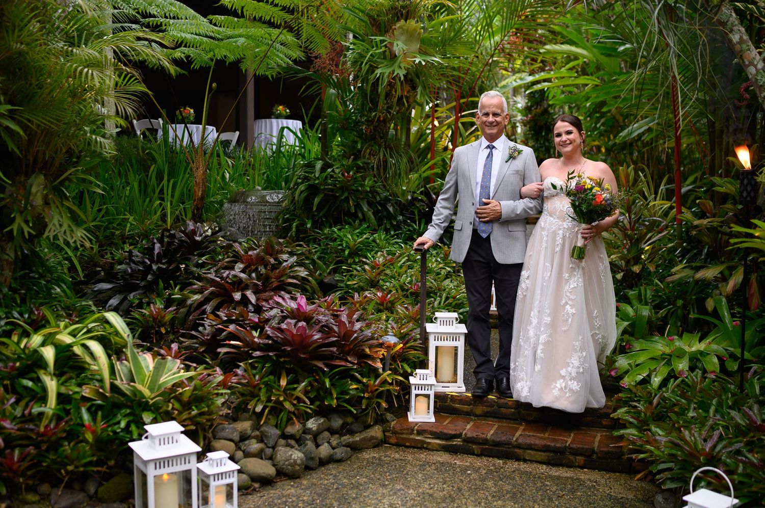 Bride and groom smiling together in a lush tropical garden wedding ceremony with lanterns and vibrant greenery.