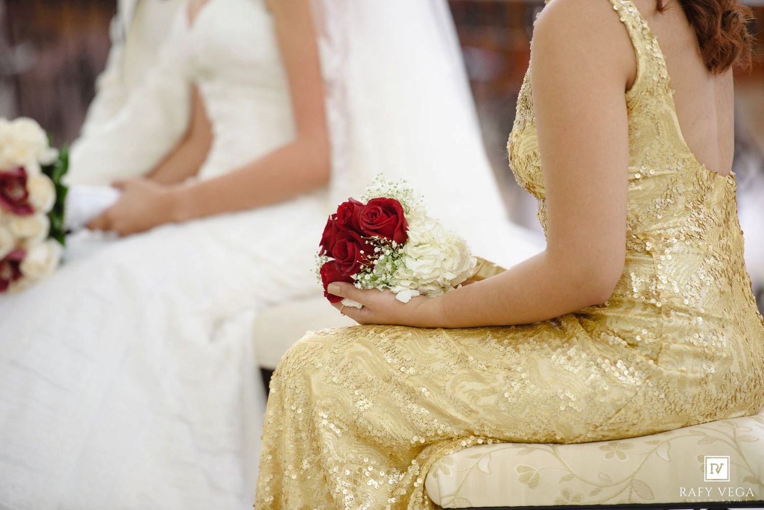 Bride in white gown and bridesmaid in gold dress holding red rose bouquet seated at a wedding ceremony.