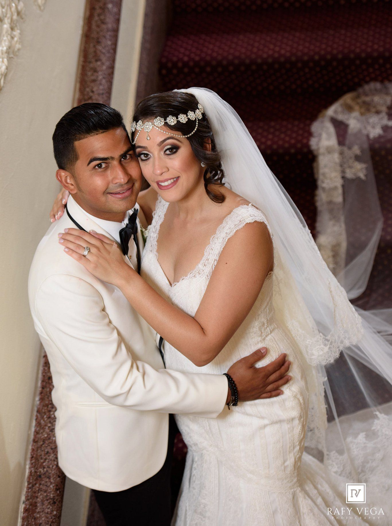 Elegant bride and groom posing together on staircase, bride wearing lace wedding gown with veil and floral headpiece.