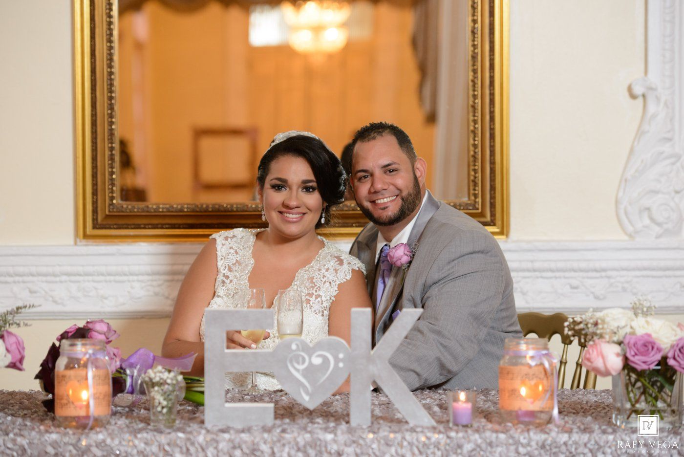 Bride and groom smile together at sweetheart table decorated with candles and monogram letters at wedding reception.