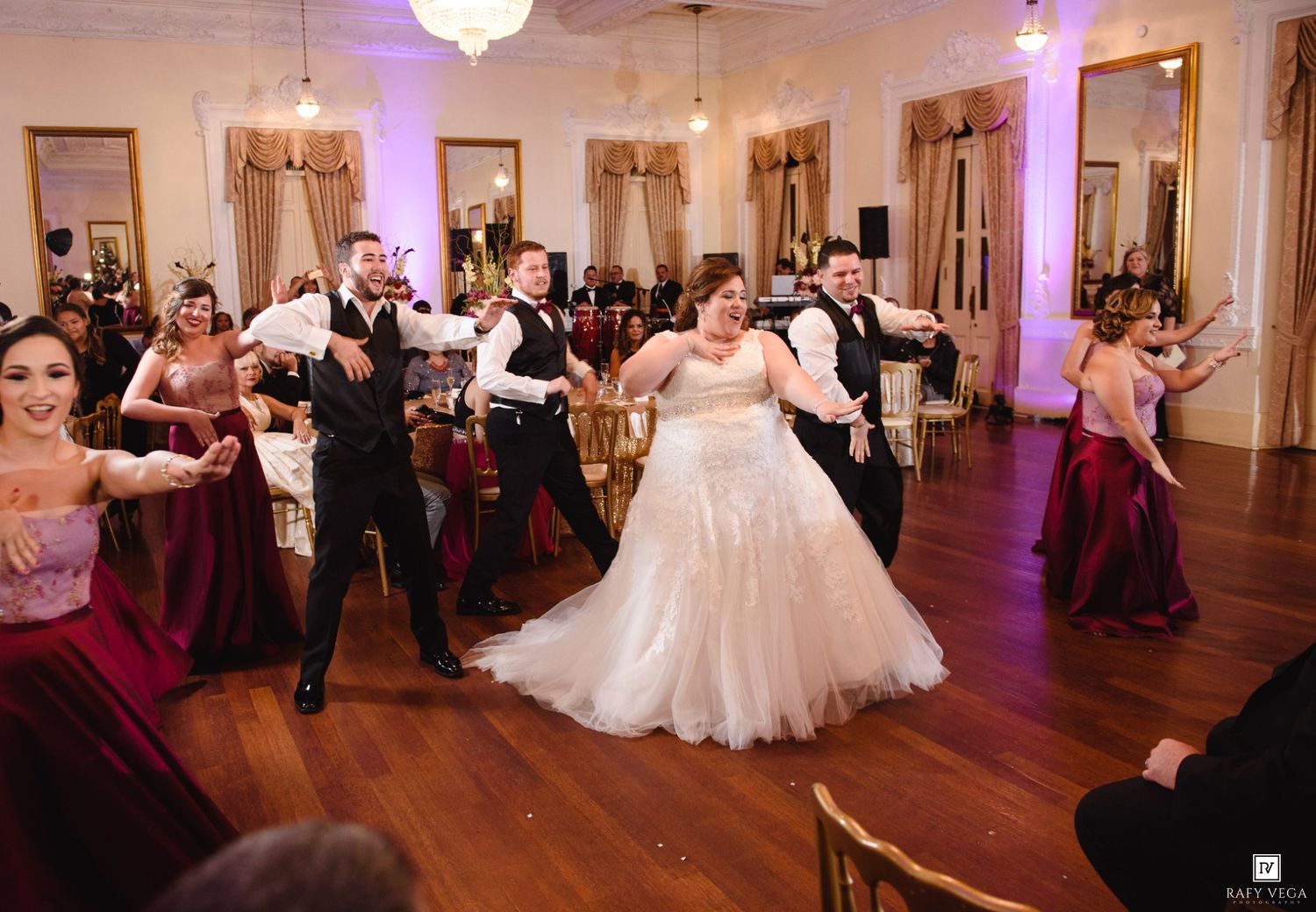 Bride in white gown dances with groomsmen and bridesmaids in burgundy dresses at elegant wedding reception.