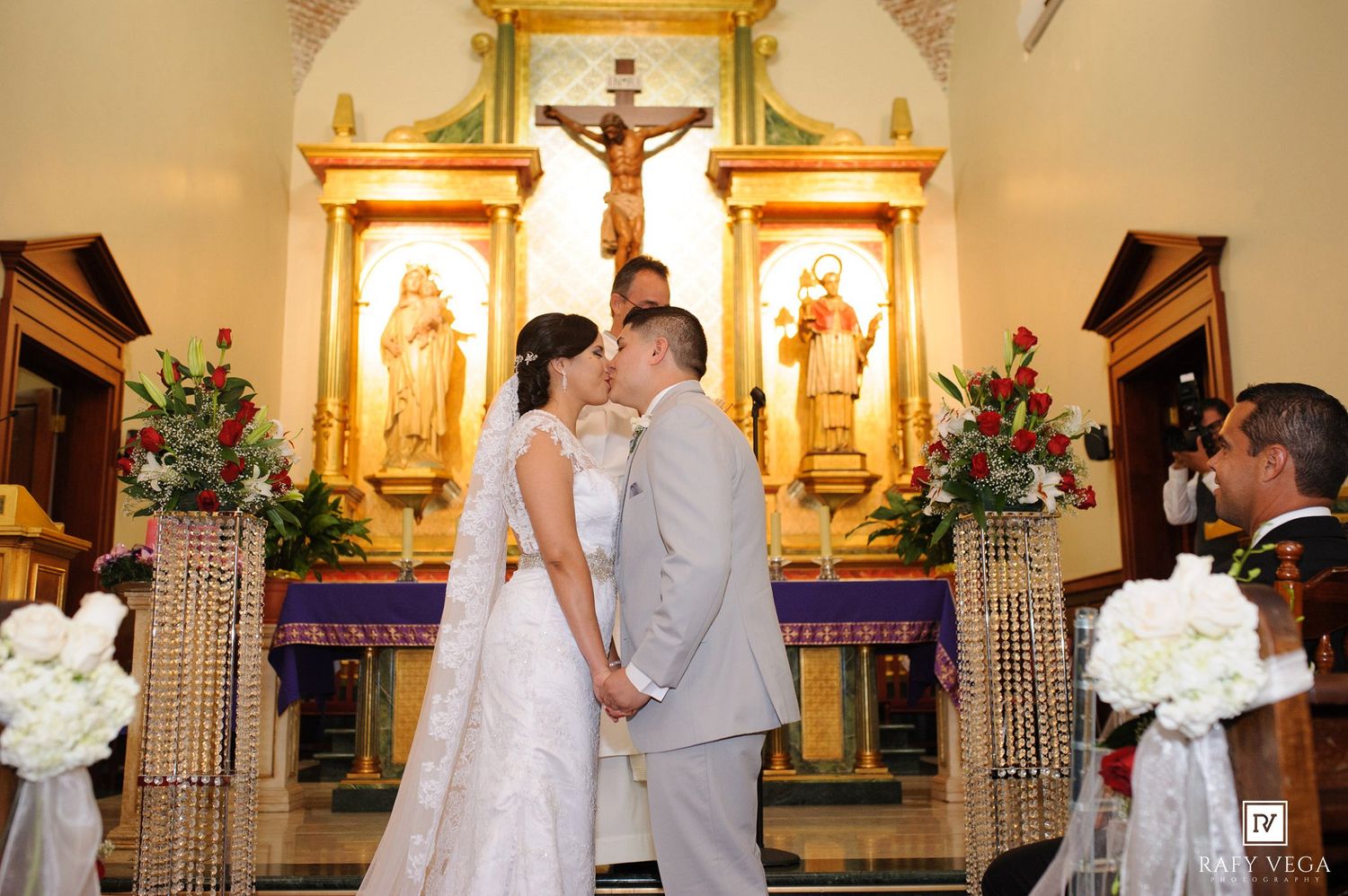 Bride and groom sharing first kiss at ornate church altar during wedding ceremony.