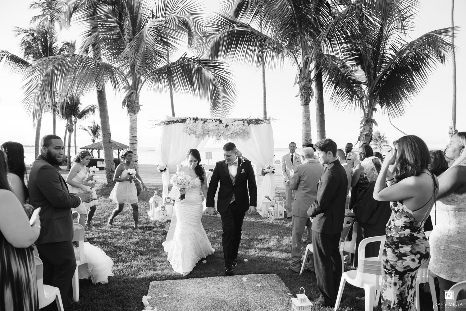 Bride and groom walk down the aisle at an outdoor beach wedding ceremony surrounded by palm trees.