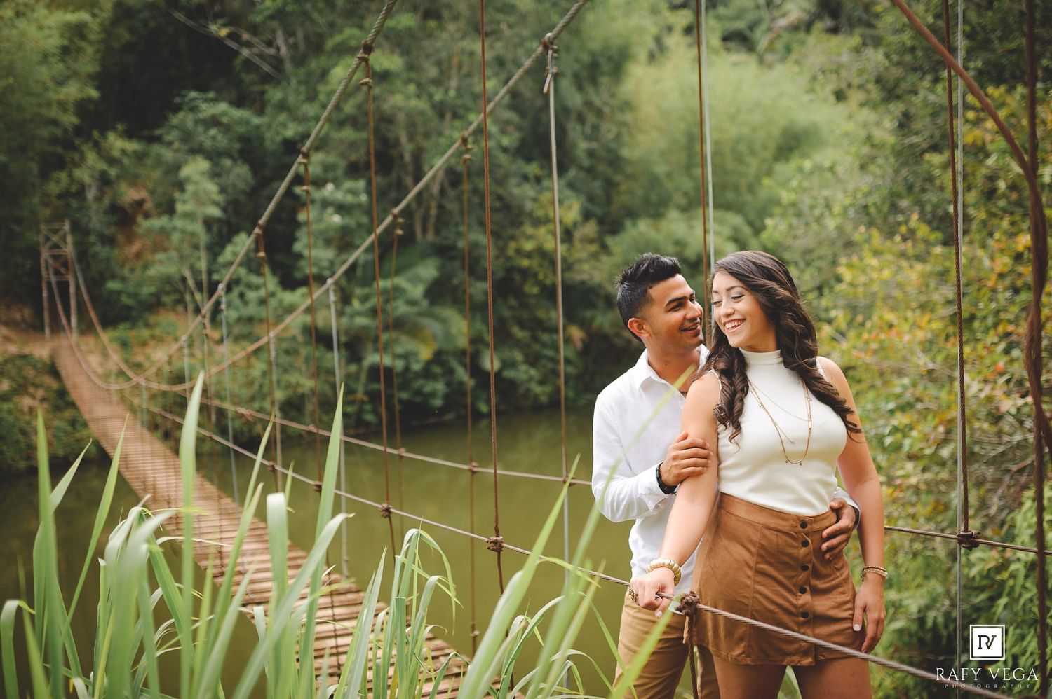 Couple smiling and embracing on a suspension bridge surrounded by lush green trees and a river backdrop.