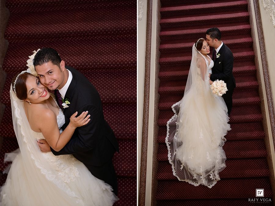Bride and groom pose on elegant red carpeted staircase, sharing a romantic kiss in formal wedding attire.