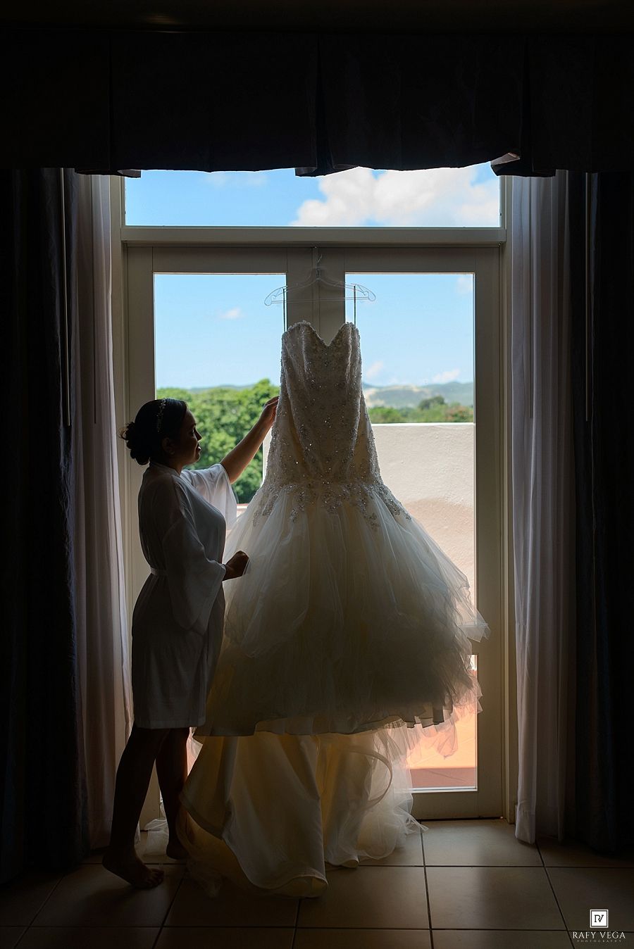 Bride admiring her elegant white wedding gown hanging in a sunlit window with a scenic outdoor view.